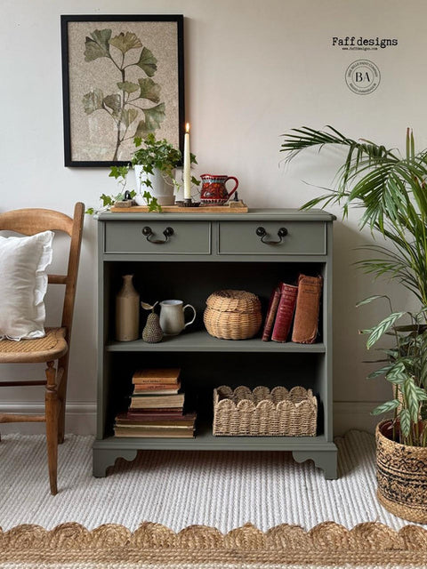A green cabinet with two drawers and open shelves holds books, baskets, and decor. A plant, candles, and a framed leaf print sit on top. A wooden chair with a white cushion is on the left. A plant in a pot is on the right, on a textured rug.