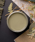 A jar filled with olive-green paint is surrounded by dried foliage and a textured fringed cloth on a dark surface. Nearby, there is a book with a green cover, adding a rustic and natural feel to the scene.