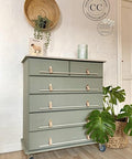 Dresser with five drawers painted with all in one mineral paint in a sage green, plant to the right in a wicker basket, basket on wall with greenery and a candle and a picture of a sheep