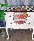A white ornate dresser stands against a textured gray and blue wall. The dresser features two drawers with floral and checkered patterns. A small potted plant is placed to the left, and part of the wall has cascading paint streaks. The carpet is beige.