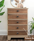 A wooden dresser with five drawers stands between two plants. On top, there's a round mirror and a decorative vase. A label that reads "Tobacco Road" points to the dresser's color.