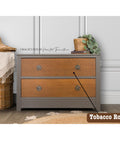 A two-drawer wooden dresser with a gray frame and natural wood front stands against a white paneled wall. A wicker basket with a fringed cloth and a small plant on top add decorative touches. A label reads "Tobacco Road" in the corner.