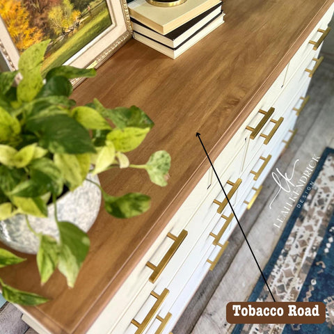 A wooden sideboard is adorned with books and a framed painting. A potted plant with variegated leaves is placed on the sideboard. The furniture features gold-accented drawer handles. The words "Tobacco Road" are on the bottom right corner.