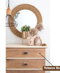 A dresser with a rustic wooden finish, topped with decorative items including a potted green plant, dried pampas grass, and a round mirror with a woven frame. A label reading "Tobacco Road" points to the dresser.