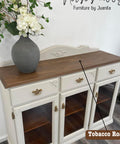 A vintage-style sideboard with a wooden top and white painted cabinets. It has three glass-front cabinets and ornate brass handles. On top, there’s a large decorative vase filled with white flowers and greenery. A label reads "Tobacco Road.