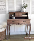 A vintage wooden desk with ornate carvings stands against a white-paneled wall. It features open shelves, small drawers, and a decorative vase on top. A mug is on the desktop. A woven basket is on the floor beside it.