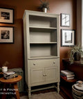 A tall, light brown open-shelf cabinet with two drawers stands in a cozy room with brown walls, framed art, plants, a chair, and a window. Text reads â€œLost &amp; Found Mercantileâ€ in the lower left corner.