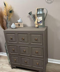 A small gray-brown dresser with gold knobs stands on a wood floor. On top are two ceramic birds, a bundle of books, a watering can with a beaded tassel, and pampas grass in a vase beside it. "Reimagined by Patty" is written in script.