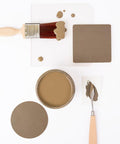 A paintbrush with brown paint, a jar filled with brown paint, a painted square, a round sample, and a palette knife with paint are arranged neatly on a white background.