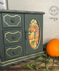 Small green jewelry cabinet with decorative orange design on a wooden surface with oranges and leaves.