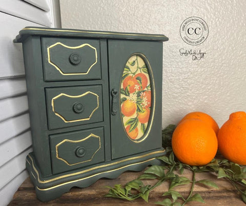 Small green jewelry cabinet with decorative orange design on a wooden surface with oranges and leaves.