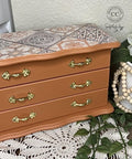 Wooden jewelry box with decorative top, gold handles, and a white vase with flowers on a lace trivet.