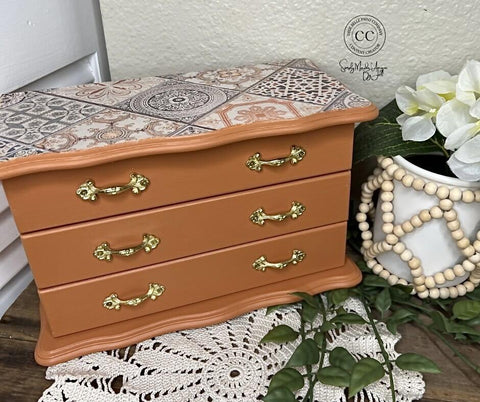 Wooden jewelry box with decorative top, gold handles, and a white vase with flowers on a lace trivet.
