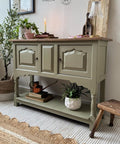 A small green sideboard with two paneled doors stands against a wall, topped with decor items, a lit candle, and books. A plant and books sit on the lower shelf; a woven rug and basket are nearby.