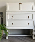 A white vintage writing desk with two drawers, brass handles, and slanted top stands against a light wall. A fern in a woven basket and a decorative sculpture are on the floor beside it. The McKAY Designs watermark is in the corner.