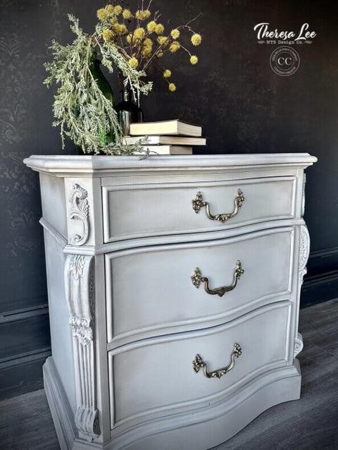 A vintage-style gray dresser with ornate brass handles stands against a dark patterned wall. A vase with yellow and green flowers, along with stacked books, adorns the top. The floor is a dark wood. The photo and piece is by Theresa Lee.