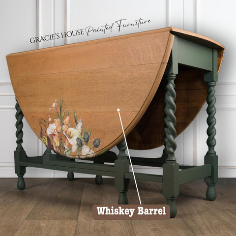 Wooden table with a foldable top, decorated with floral designs and labeled "Whiskey Barrel." Finished with a water-based stain, it boasts dark green, twisted legs. Text at the top reads "Gracie's House Painted Furniture," set against a white paneled wall.