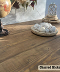 A wooden table displays a globe on a stand, a vase with flowers, and a wooden tray of decorative white balls. Sunlight dances across the surface, revealing the rich, water-based stain of Charred Hickory etched at the bottom right.