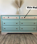 A dresser with six teal drawers and two light wood drawers on top. There's a vase with pampas grass on top. An arrow points to one of the light wood drawers labeled "White Magic." The floor is wooden.
