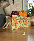 A decorative box with orange and green floral patterns with oranges is filled with grapes and oranges, placed on a wooden countertop. A rope handle is on the side of the box. Kitchen items and a window are in the background.