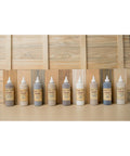 Nine bottles of Dixie Belle VooDoo Stains in varying shades are lined up on a table. The background features wooden planks in different natural tones, complementing the stains.