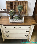 A vintage dresser with a dark walnut top and cream-colored body. It has ornate drawer handles. On top, there's a decorative tray holding two small bush sculptures and a circular metal decor piece. A "Creative Moments" watermark is at the bottom.