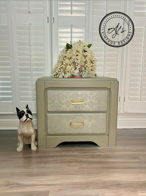 A mute green nightstand with Wedding Lace Transfer seen applied to the fronts of the drawer has gold hardware. Staged next to a wooden black and white dog statue and a short vase with large white flowers cover majority of it on top of the nightstand.