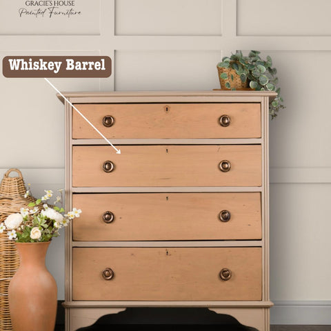 A wooden dresser with four drawers, labeled "Whiskey Barrel," stands against a paneled wall. A wicker basket with flowers and a small plant decorate the scene. The dresser is under watermark reading "Gracie's House Painted Furniture.