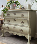 A vintage-style sage green dresser with ornate gold handles stands against a white wall, decorated with potted plants, a mug, a tray, and a picture frame on top. The dresser features curved legs and intricate detailing.