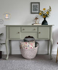 A pale green vintage side table with three drawers is against a white wall. A vase with yellow flowers, a framed picture, and a lantern sit on top. A basket with floral pillows is underneath; a pink chair is on the right.