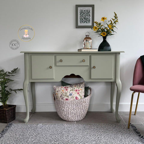 A pale green vintage side table with three drawers is against a white wall. A vase with yellow flowers, a framed picture, and a lantern sit on top. A basket with floral pillows is underneath; a pink chair is on the right.