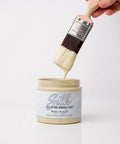 A hand holds a paintbrush dipped in light green paint above an open jar labeled "Silk All-In-One Mineral Paint, Wicklow Glen." Paint is dripping from the brush into the jar against a white background.