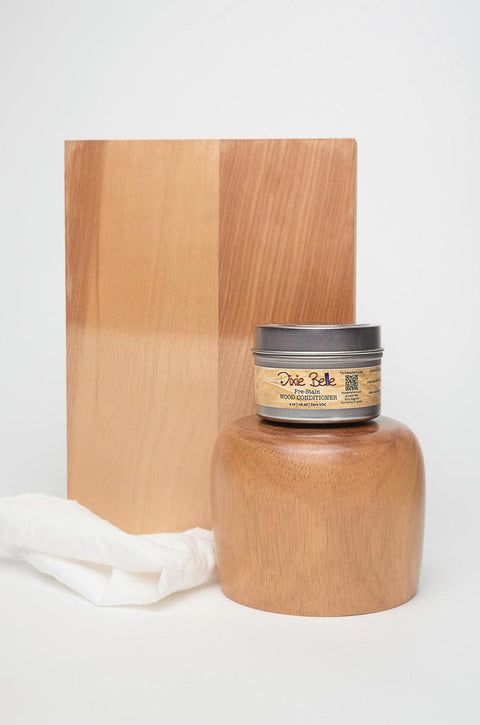 A jar of Dixie Belle wood conditioner is placed in front of a wooden block and a polished wooden object. A white cloth rests beside the jar, all set against a plain white background.