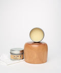 An open jar of Dixie Belle wood conditioner is placed in front of a wooden block and a polished wooden object. A white cloth rests beside the jar, all set against a plain white background.
