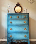 Applique #2152 Set of 2 is the perfect mouldable embellishment for any project!
A blue chest of drawers with gold hardware and at the bottom trim is seen Applique #2152 painted in silver. Staged on top of the piece is a bronze framed mirror.