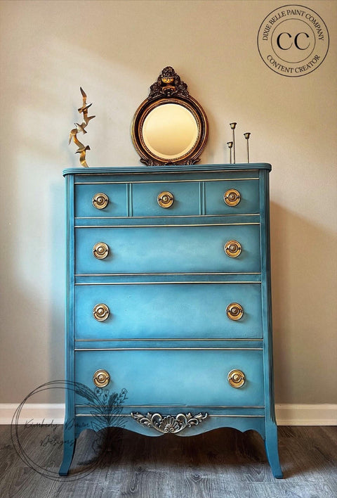 Applique #2152 Set of 2 is the perfect mouldable embellishment for any project!
A blue chest of drawers with gold hardware and at the bottom trim is seen Applique #2152 painted in silver. Staged on top of the piece is a bronze framed mirror.