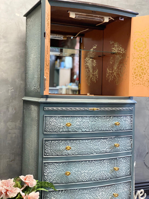 WoodUBend Trim #TR50a is the perfect mouldable embellishment for any project!  A blue chalk painted China cabinet with gold knobs and a white chalk painted stenciled pattern on the side and drawers.  The inside of the cabinet is chalk painted orange with a gold stenciled pattern and a mirror in the back.  Staged against a gray wall with pink flowers and greenery sitting next to the cabinet.