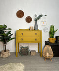 Yellow painted two drawer dresser transformation with bronze pulls. Staged with a gray textured vase containing faux greenery and a bamboo candle holder on top.