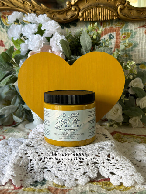 Yellow painted wooden heart with a jar of Yellowstone in front of it. Staged on crocheted lace with faux flowers and greenery in the background.